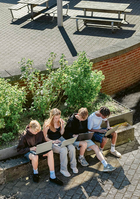 Four people sitting outdoors on a low concrete wall with laptops, surrounded by greenery and brick walls, working or studying together in casual clothing. Description generated by AI