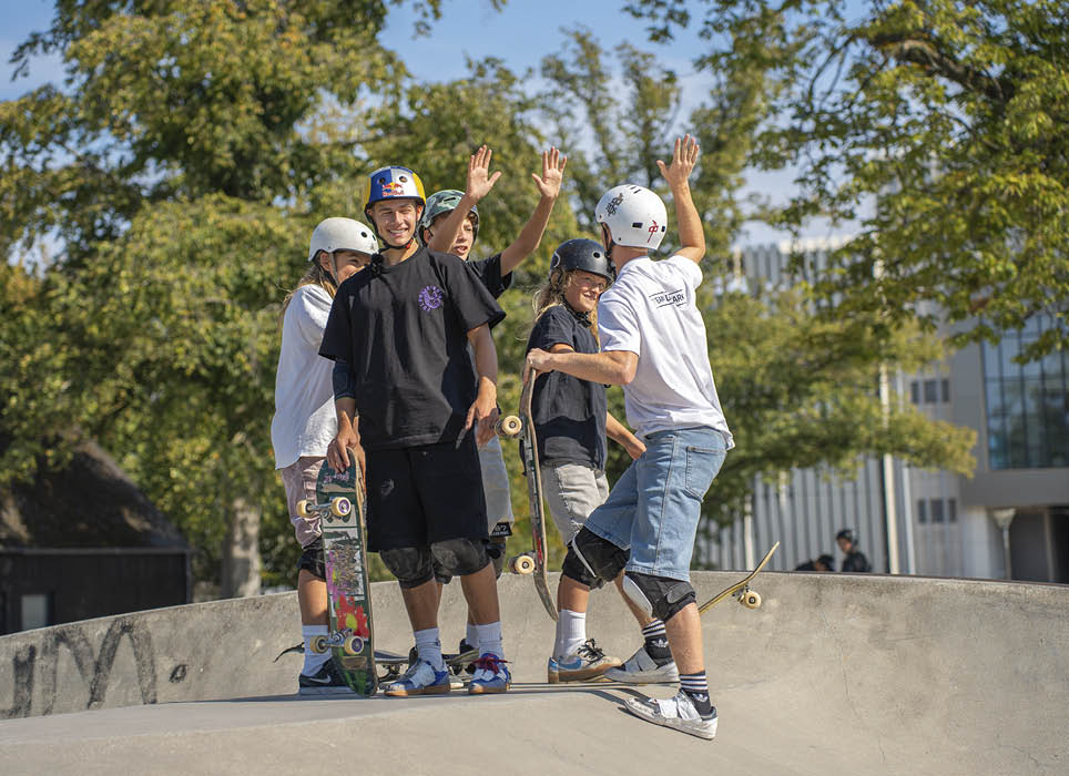 A group of people wearing casual clothing and protective gear are standing with skateboards at a skate park on a sunny day. Description generated by AI