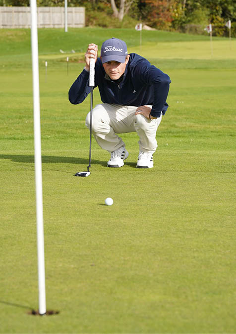 A person wearing a navy blue jacket, beige pants, white sneakers, and a cap is crouching on a golf course while holding a golf club. Description generated by AI
