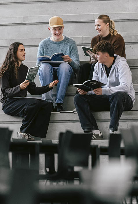 Four people sitting on concrete steps indoors are smiling and reading books together in a casual setting. Description generated by AI