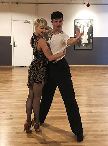 Two adults dressed in dance attire are posing closely together in a dance studio with wooden floors and a framed photo on the wall behind them. Description generated by AI